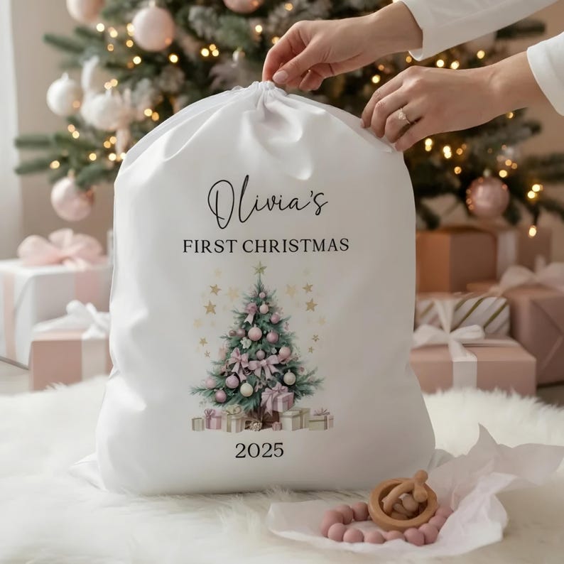 May include: A white Christmas gift bag with a drawstring closure. The bag features the text "Olivia's FIRST CHRISTMAS" above a Christmas tree design with pink ornaments and bows. The year "2025" is printed below the tree.