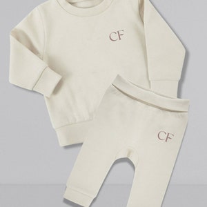 May include: A cream colored baby outfit set with a long-sleeved sweatshirt and matching pants. Both pieces feature the initials "CF" embroidered in a light pink color.