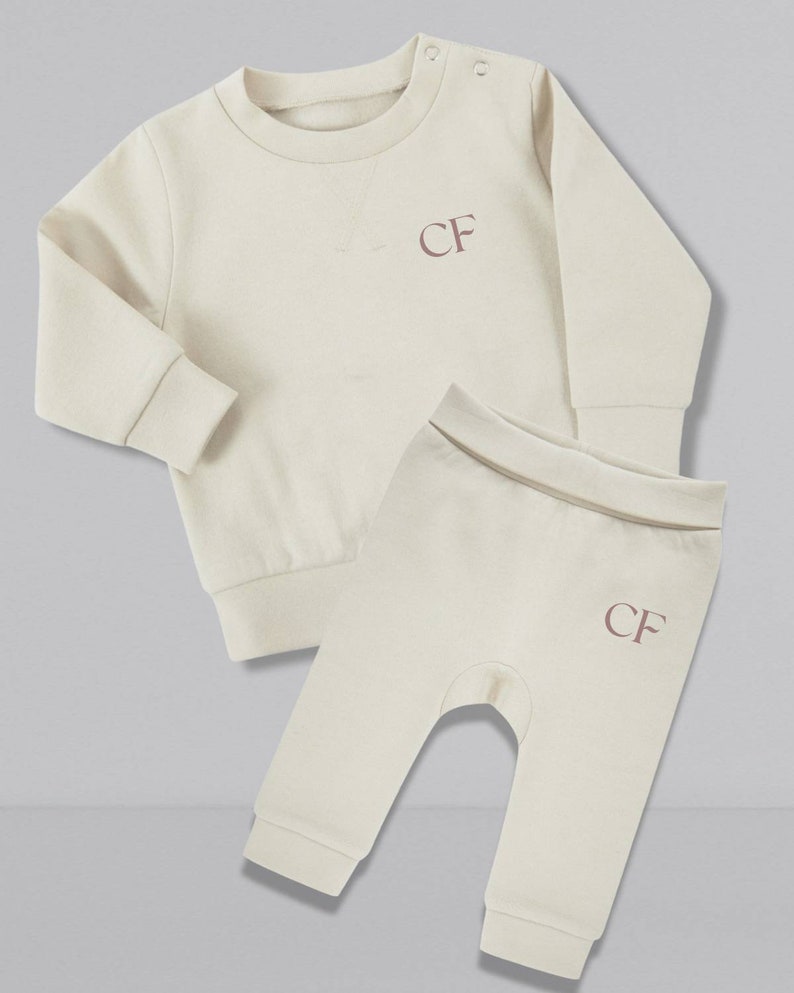 May include: A cream colored baby outfit set with a long-sleeved sweatshirt and matching pants. Both pieces feature the initials "CF" embroidered in a light pink color.