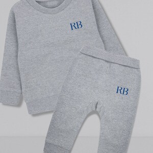 May include: A gray two-piece baby outfit with long sleeves and pants. The top has a round neckline and the pants have a cuffed bottom. Both pieces have the initials "RB" embroidered in blue on the front.