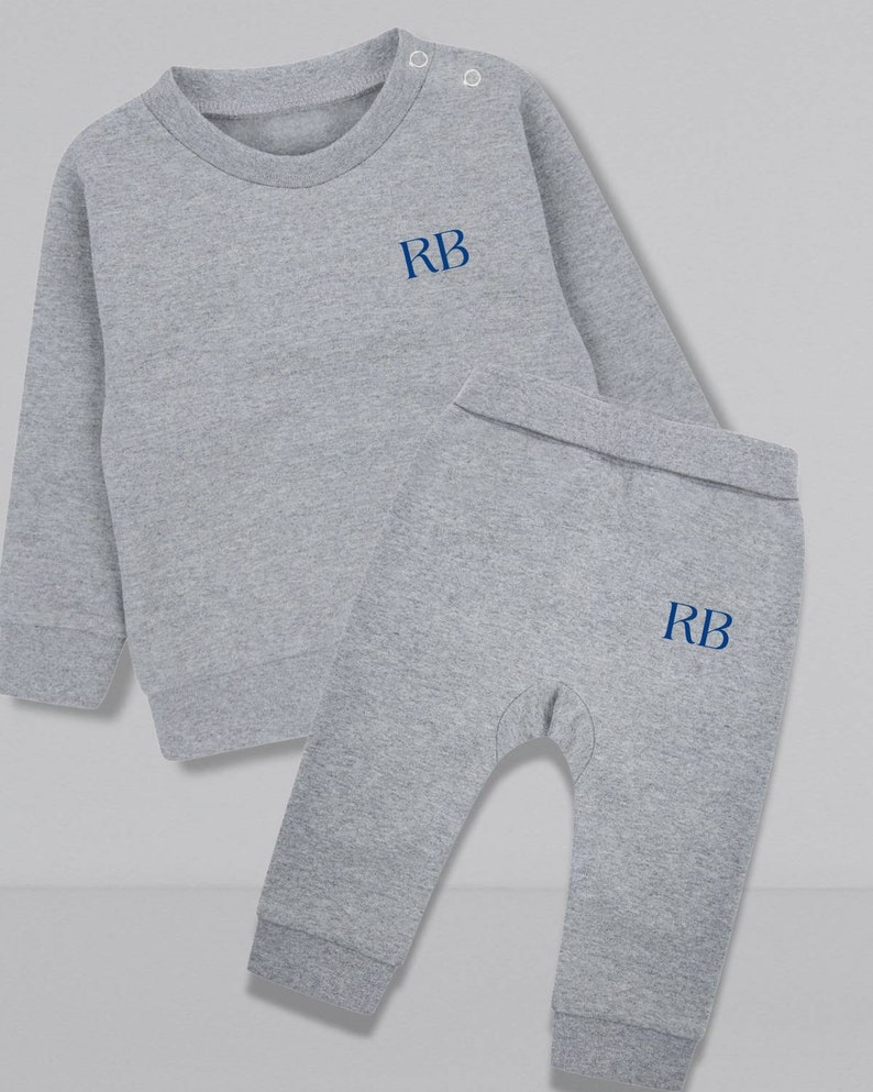 May include: A gray two-piece baby outfit with long sleeves and pants. The top has a round neckline and the pants have a cuffed bottom. Both pieces have the initials "RB" embroidered in blue on the front.
