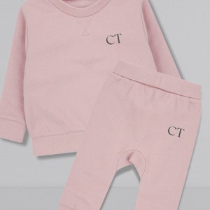 May include: A pink baby outfit set with a long-sleeved sweatshirt and matching pants. Both pieces feature the initials "CT" embroidered on the front.