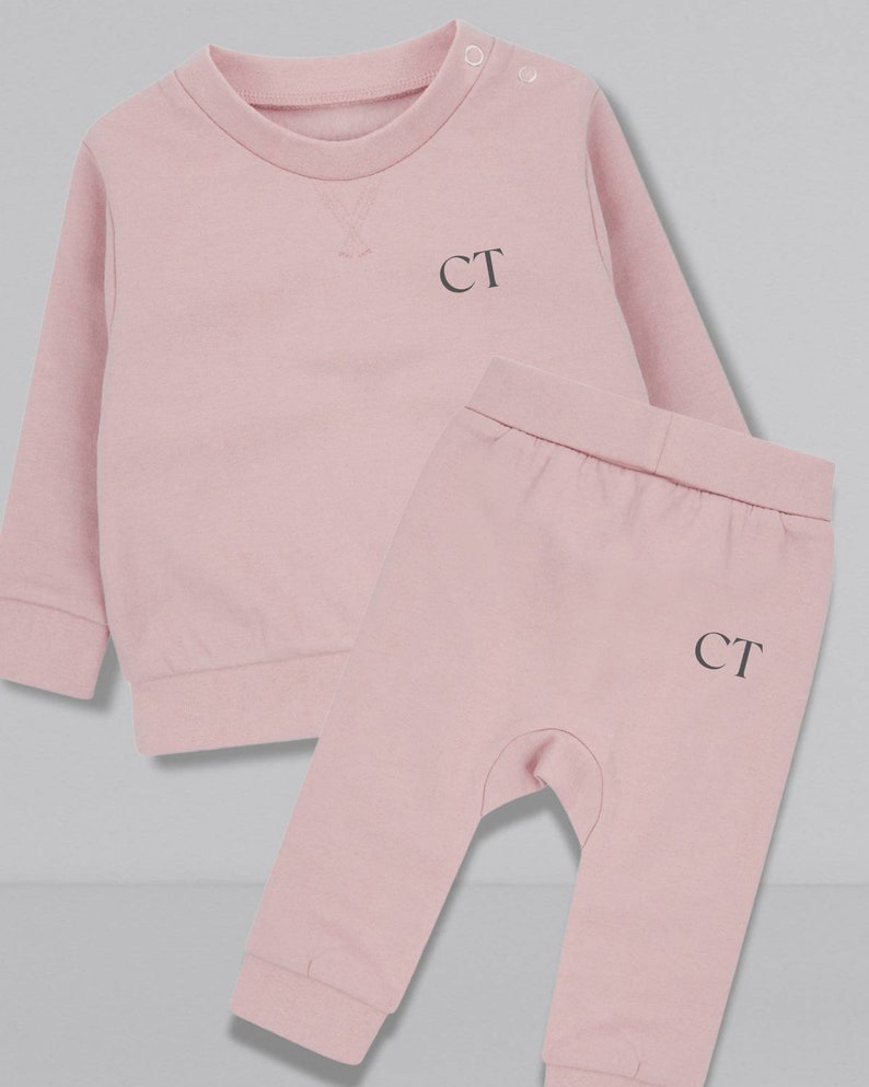 May include: A pink baby outfit set with a long-sleeved sweatshirt and matching pants. Both pieces feature the initials "CT" embroidered on the front.