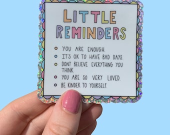 May include: A square sticker with a scalloped, holographic border featuring the text LITTLE REMINDERS in pastel colors. The list includes: YOU ARE ENOUGH, IT'S OK TO HAVE BAD DAYS, DONT BELIEVE EVERYTHING YOU THINK, YOU ARE SO VERY LOVED, BE KINDER TO YOURSELF.