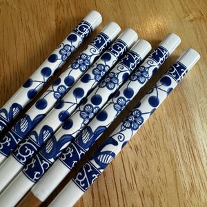 May include: Set of five white chopsticks with blue floral designs. The chopsticks are made of ceramic and have a wooden base.