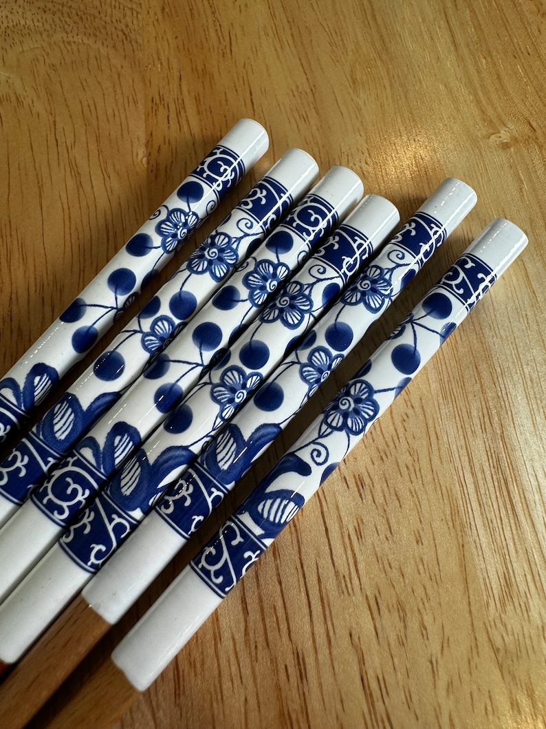 May include: Set of five white chopsticks with blue floral designs. The chopsticks are made of ceramic and have a wooden base.