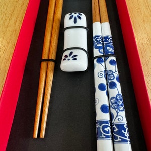May include: A set of chopsticks in a red box. The chopsticks are made of wood and have a white porcelain design with blue floral patterns. The set includes two pairs of chopsticks and two porcelain rests.