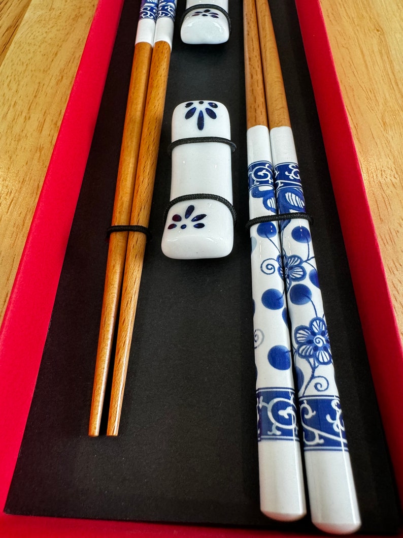 May include: A set of chopsticks in a red box. The chopsticks are made of wood and have a white porcelain design with blue floral patterns. The set includes two pairs of chopsticks and two porcelain rests.