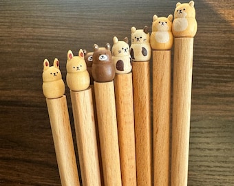 Puede incluir: Una colección de siete lápices de madera clara, cada uno rematado con una pequeña figura de animal de madera pintada a mano. Las figuras incluyen varios osos, gatos y conejos con rasgos faciales y marcas distintivas en tonos marrón, beige y blanco.