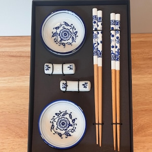 May include: A boxed set of Japanese-style tableware. The set includes two small white and blue floral patterned dishes, two pairs of chopsticks, and two chopstick rests. The chopsticks are light brown with blue floral designs.