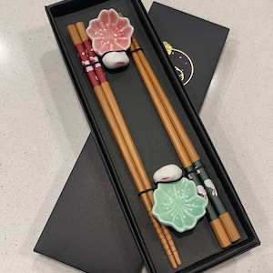 Könnte beinhalten: Ein Set mit zwei Paar Essstäbchen in einer schwarzen Box. Die Essstäbchen sind aus Bambus und haben ein Blumenmuster. Die Box enthält auch zwei kleine Keramikteller mit Blumenmustern.