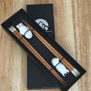 May include: A black box with a silver logo containing two wooden chopsticks with white ceramic bear-shaped rests.