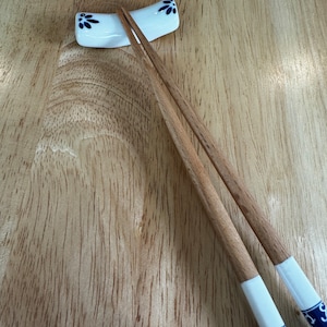 May include: A pair of wooden chopsticks with white tips and blue floral designs rest in a white ceramic chopstick holder on a wooden surface.
