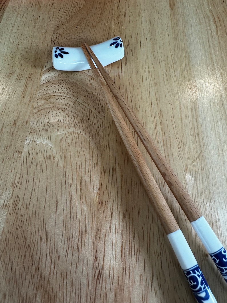 May include: A pair of wooden chopsticks with white tips and blue floral designs rest in a white ceramic chopstick holder on a wooden surface.