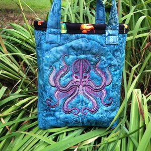 May include: A blue quilted tote bag with a black trim and an embroidered octopus design. The octopus is purple and has eight tentacles.