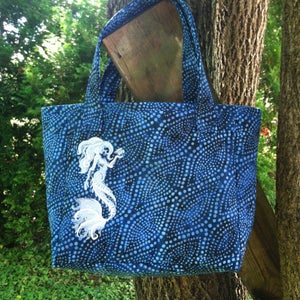 May include: Blue tote bag with a white embroidered mermaid design. The bag features a blue dotted pattern and sturdy handles. The bag is hanging from a tree branch.