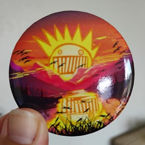May include: A round button with a sunset design. The button features a stylized sun with a face, mountains, water, and birds in flight. The color palette includes orange, yellow, purple, and black.
