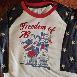 May include: A white and red raglan-style t-shirt with blue sleeves featuring yellow stars. The shirt displays the text "Freedom of '76" above a graphic of three figures with drums and a flag.