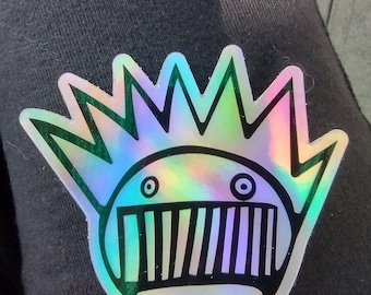 May include: A holographic sticker featuring a stylized circular face with a spiky crown-like top, two small circular eyes, and a wide mouth filled with vertical black lines, all outlined in a bold black border against a reflective rainbow surface.