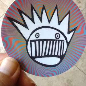 May include: A white sticker with a black and white cartoon character with a spiky hairdo and a mouth with teeth. The background is a swirling pattern of red, orange, blue, and green.