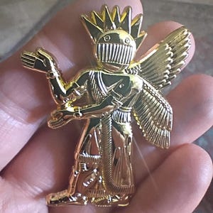May include: A shiny gold-toned metal pin shaped like a stylised winged figure. The figure features a helmet with a vertical grill, a spiked crown, and detailed wings. The figure is depicted in a standing position with one arm raised and one arm bent.