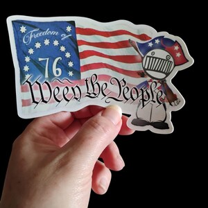May include: A white sticker with a cartoon illustration of a person holding a gun and wearing a hat with stars. The sticker features a red, white, and blue American flag with the words "Freedom 76 We the People" in a cursive font.