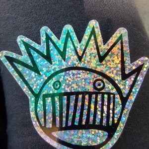 May include: A holographic sticker featuring a circular character with a spiky crown and a wide, toothy grin. The design is outlined in black, and the surface has a reflective, multi-colored glitter effect that shifts in the light.