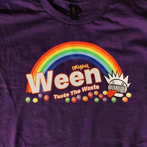 Könnte beinhalten: Ein lila T-Shirt mit einem Regenbogen über dem Wort "Ween" in Weiß und dem Text "Taste The Waste" darunter. Rechts befindet sich eine Cartoonfigur mit Krone und Zunge. Bunte Punkte sind darunter verstreut.