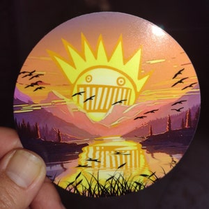 May include: A round sticker with a sunset scene featuring a stylized yellow sun with a toothy grin and a mountain range in the background. The sticker has a glossy finish.