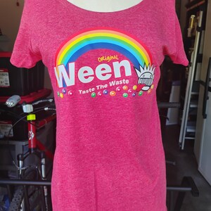 May include: A heathered pink short-sleeve t-shirt featuring a graphic of a rainbow above the white text "Ween" and "Taste The Waste". The design includes a stylised character with a spiky head and tongue out, surrounded by small colourful circles.
