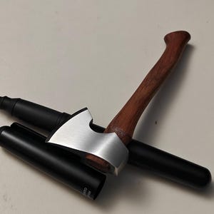 Pode incluir: Uma caneta preta com um porta-caneta prateado em forma de machado e um cabo de madeira. A caneta está sobre uma superfície clara. O porta-caneta tem uma cabeça de machado prateada e um cabo de madeira castanha.