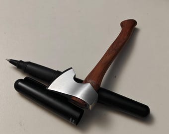 Pode incluir: Uma caneta preta com um porta-caneta prateado em forma de machado e um cabo de madeira. A caneta está sobre uma superfície clara. O porta-caneta tem uma cabeça de machado prateada e um cabo de madeira castanha.