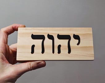 May include: A rectangular wooden sign with the Hebrew word "יהוה" cut out in black. The sign is made of light-colored wood and is held in a hand.