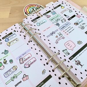 May include: A pink planner with black polka dots and colorful stickers. The stickers feature illustrations of a watering can, a phone, a shopping basket, a glass bin, and a reminder to attend a big event next month. The planner has a tab that says "Today".