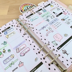 May include: A pink planner with black polka dots and colorful stickers. The stickers feature illustrations of a watering can, a phone, a shopping basket, a glass bin, and a whale. The planner has a tab that says "Today".