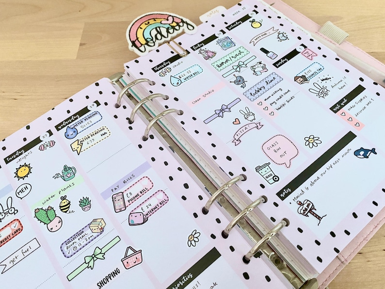 May include: A pink planner with black polka dots and colorful stickers. The stickers feature illustrations of a watering can, a phone, a shopping basket, a glass bin, and a whale. The planner has a tab that says "Today".