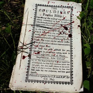 May include: An open, antique book titled "The Souldiers Pocket Bible" with blood splatters on the pages. The book is printed in black ink with decorative borders and text from 1643. The book is lying on a bed of green grass.