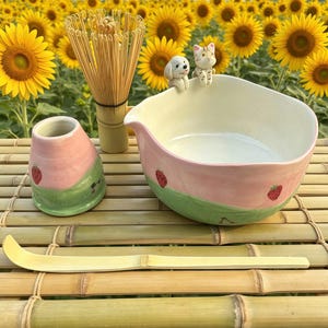Op de afbeelding: Een matcha theeset met een roze en groene kom, een klopper, een lepel en een keramische houder. De kom is versierd met aardbeien en kleine beeldjes van een hond en een kat. De set is geplaatst op een bamboe oppervlak.