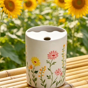 May include: A cylindrical, off-white ceramic vase with a floral design featuring yellow, pink, and orange flowers. The vase has a wavy top and a single hole. The background is a field of sunflowers.