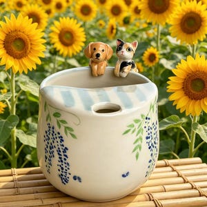 May include: A ceramic planter with a light blue and white striped rim, featuring a dog and cat figurine. The planter is decorated with blue floral designs and sits on a bamboo surface, with a backdrop of sunflowers.