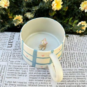 Puede incluir: Una taza de cerámica con un diseño azul claro y blanco, con una pequeña figura de gato en el interior. La taza tiene un asa y está decorada con rayas azules y pequeños corazones rosas. El fondo incluye flores amarillas.