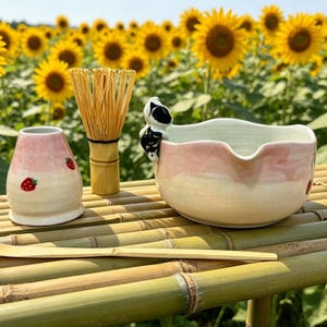 May include: A matcha tea set with a pink and white bowl, a bamboo whisk, a strawberry-decorated ceramic container, and a bamboo scoop. The set is arranged on a bamboo table with a backdrop of sunflowers.