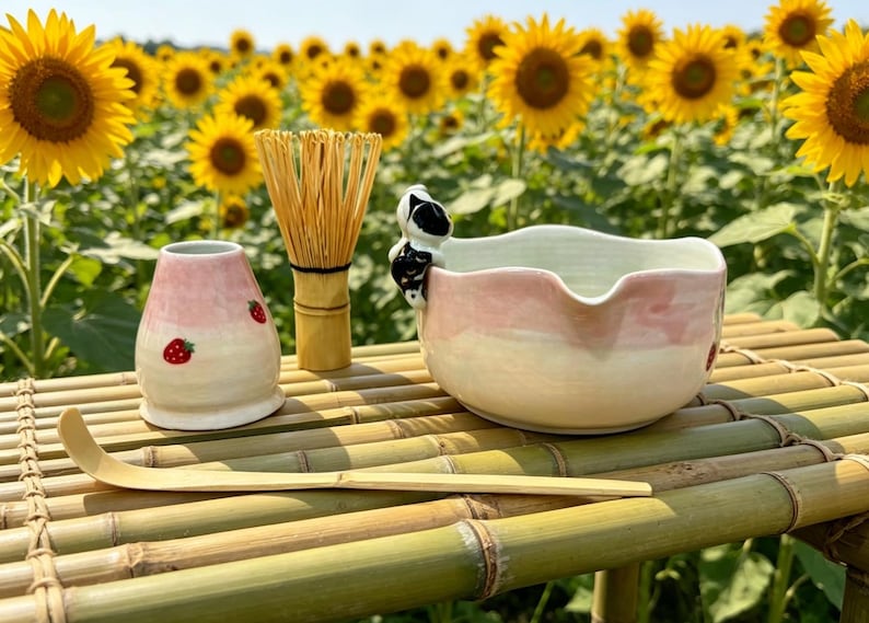 May include: A matcha tea set with a pink and white bowl, a bamboo whisk, a strawberry-decorated ceramic container, and a bamboo scoop. The set is arranged on a bamboo table with a backdrop of sunflowers.