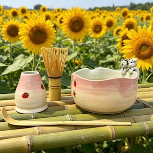 May include: A tea ceremony set with a pink and white bowl, a strawberry-decorated container, a bamboo whisk, and a scoop. The set is on a bamboo surface, with a field of sunflowers in the background.