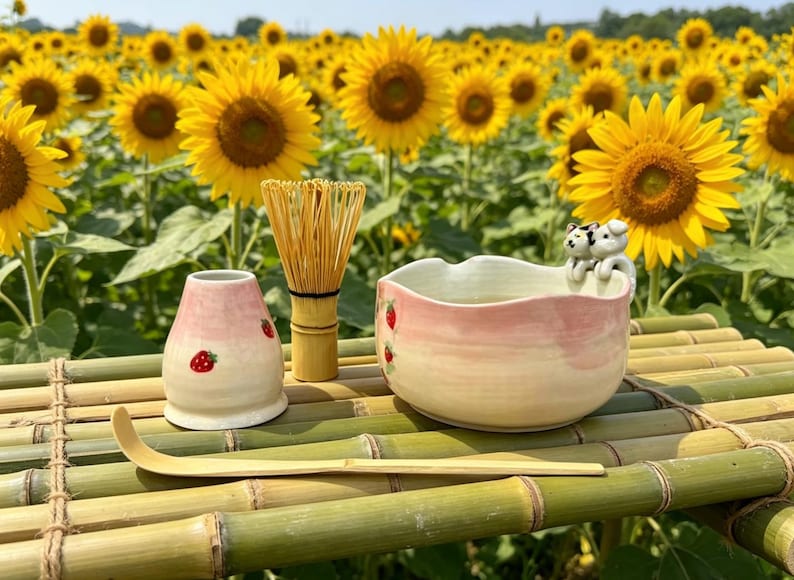 May include: A tea ceremony set with a pink and white bowl, a strawberry-decorated container, a bamboo whisk, and a scoop. The set is on a bamboo surface, with a field of sunflowers in the background.
