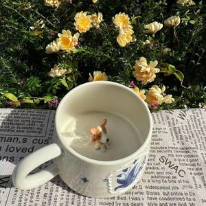 Puede incluir: Taza de cerámica blanca con una pequeña figura de perro en su interior. La taza tiene un diseño floral azul y un asa grande. El fondo presenta flores amarillas y una impresión de estilo periódico.