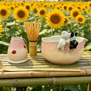 May include: A tea ceremony set with a pink and white ceramic bowl, a bamboo whisk, a strawberry-decorated container, and a bamboo scoop. The bowl has two small figurines. The set is on a bamboo table with a sunflower field in the background.
