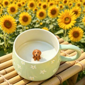 Puede incluir: Una taza de cerámica verde claro con interior blanco y una pequeña figura de perro salchicha marrón en su interior. La taza está decorada con flores de margarita blancas. El fondo presenta un campo de girasoles amarillos.