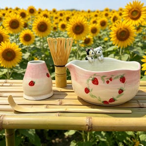 May include: A ceramic matcha bowl, whisk, and accessories are arranged on a bamboo surface. The bowl is decorated with a strawberry design. A pink and white ceramic container with strawberry accents sits next to a bamboo whisk. A wooden scoop is also present.
