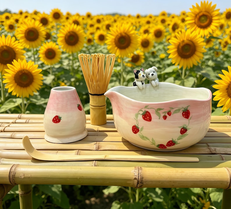 May include: A ceramic matcha bowl, whisk, and accessories are arranged on a bamboo surface. The bowl is decorated with a strawberry design. A pink and white ceramic container with strawberry accents sits next to a bamboo whisk. A wooden scoop is also present.